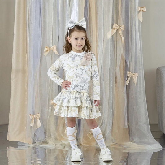 A young girl stands in front of a decorative backdrop of sheer beige and gray tulle curtains with ribbon bows, wearing a long-sleeve white dress with a light beige bunny pattern, a tiered ruffled skirt, white knee-high socks, and white sneakers.